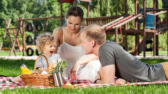 Familie beim Picknick auf Wiese mit Spielplatz im Hintergrund | Quelle: © Photographee.eu / Adobe Stock