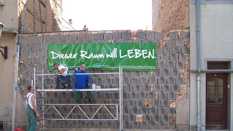 Bauarbeiten an einer Wand, an der ein Banner hängt mit der Aufschrift „Dieser Raum will LEBEN“. | Quelle: © Claus-Peter Neumann, DSK